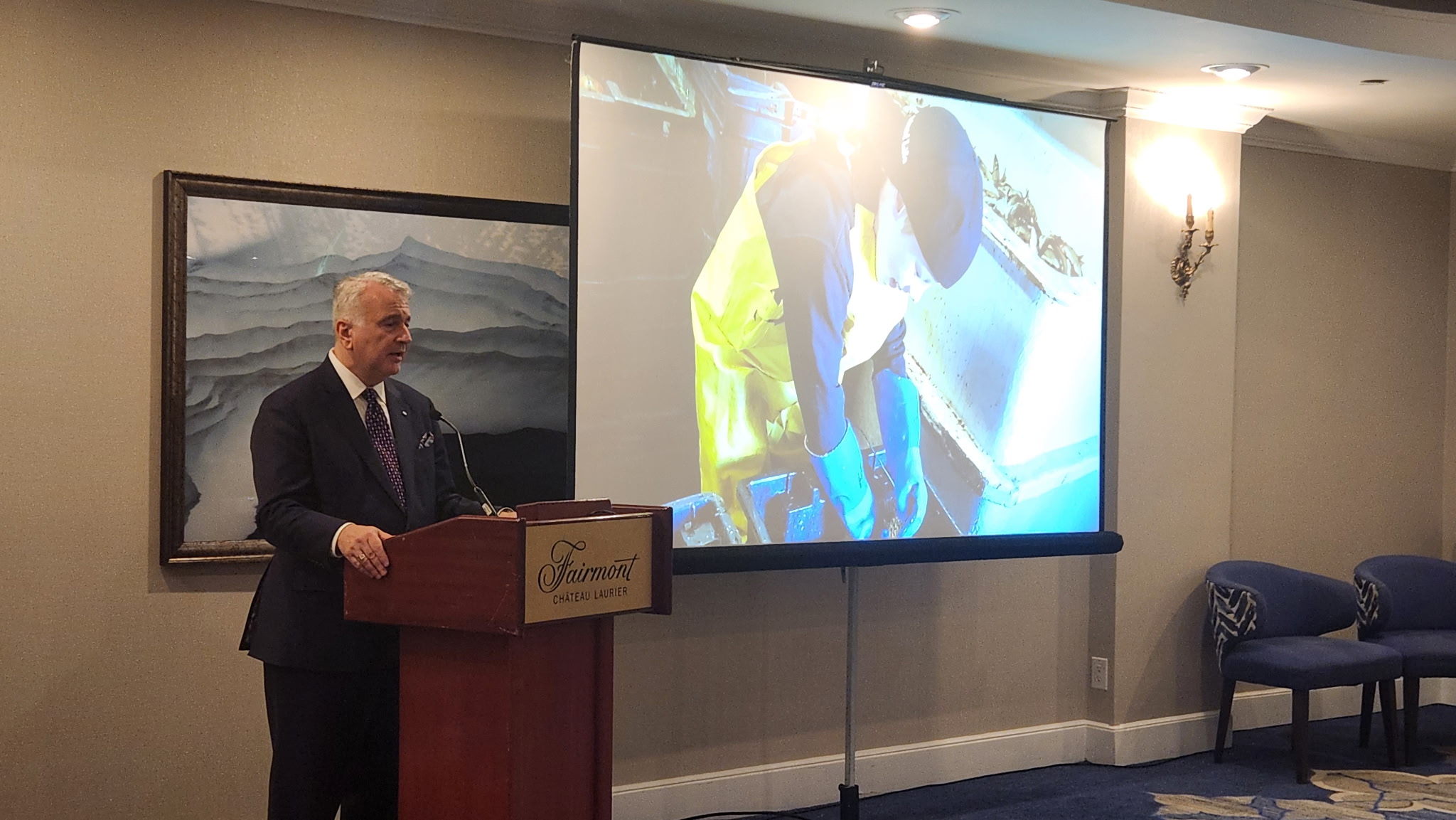 Speaker at a podium with a screen behind showing a harvester at work on deck