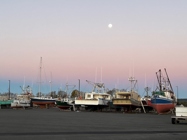 Fishing boats on drydock at sunset with moon visible against a beautiful pink and blue sky