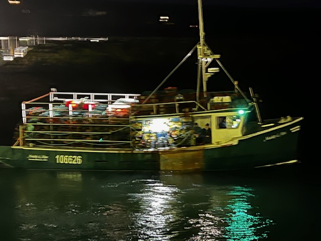 Fishing vessel number 106626 with green hull loaded with lobster traps, heading out to sea at night